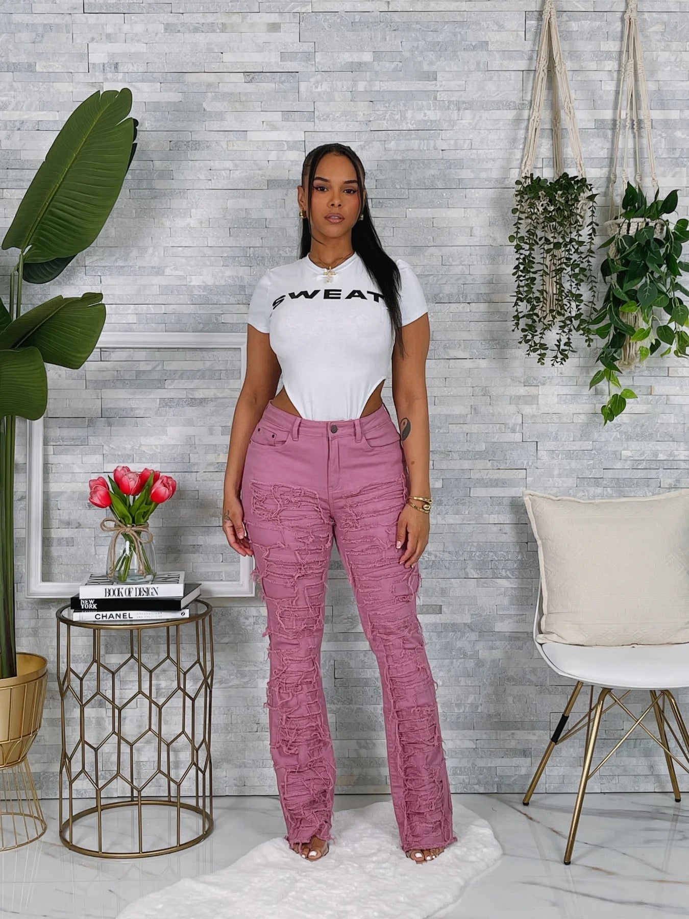 woman wearing a white t-shirt with text and pink pants standing in a room with plants and decor.