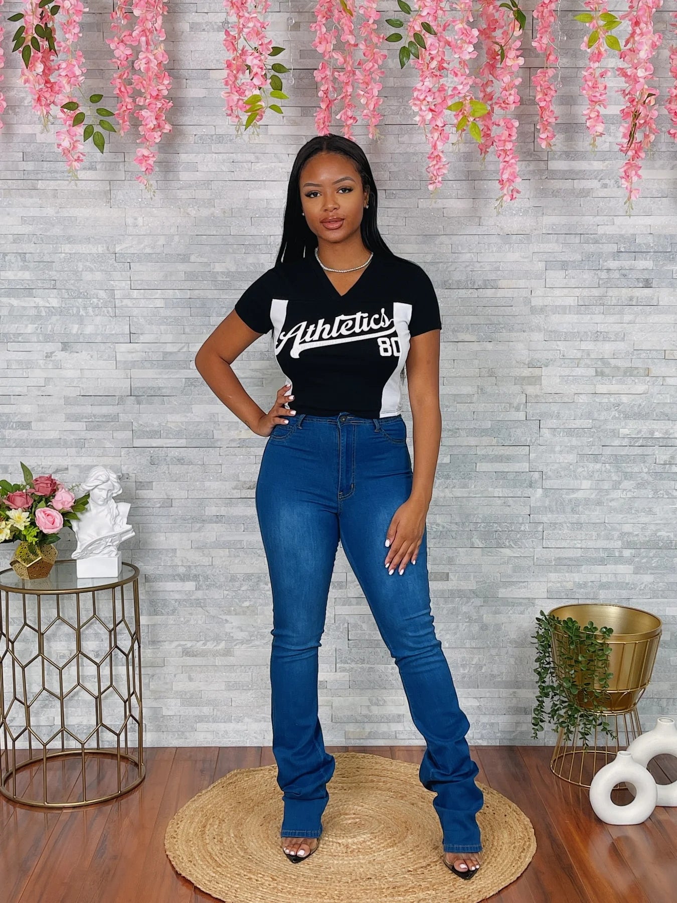 woman wearing a black t-shirt with white text and blue jeans standing in front of a decorative wall with pink flowers.